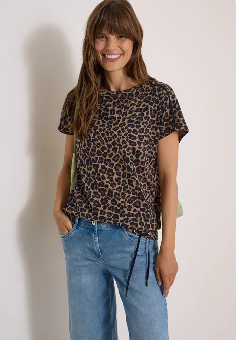 Frau mit langen Haaren lächelt, trägt Leopardenshirt und blaue Jeans vor hellem Hintergrund.