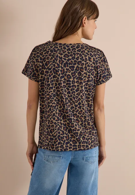 Frau steht Rücken zur Kamera, trägt kurzärmeliges T-Shirt mit Leopardenmuster und blaue Jeans vor beigem Hintergrund.