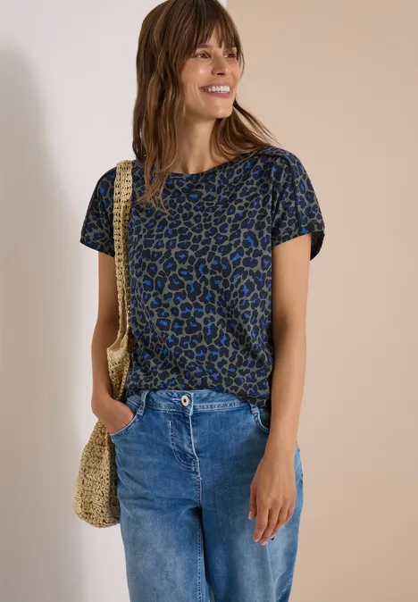 Frau in blauem Leo-Shirt, Jeans und Strohtasche lächelnd vor neutralem Hintergrund.