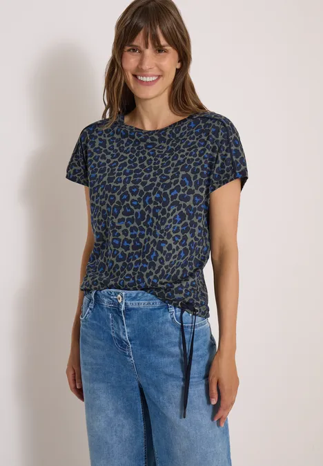 Frau in blau-schwarz gemustertem T-Shirt mit Seitenschnürung, kombiniert mit blauen Jeans, lächelt in die Kamera.