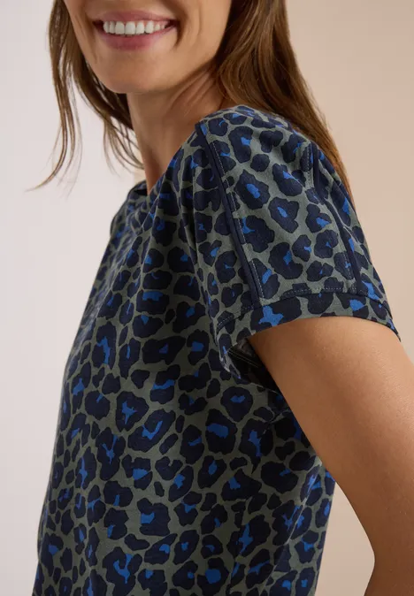 Frau in blau-grauem Leopardenmuster-Shirt lächelt seitlich vor beigem Hintergrund.