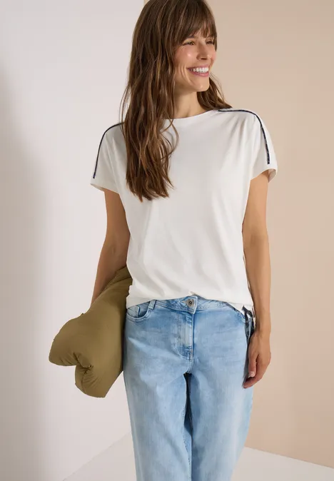 Vrouw glimlacht, draagt een wit T-shirt en lichtblauwe jeans en houdt een groen kussen vast.