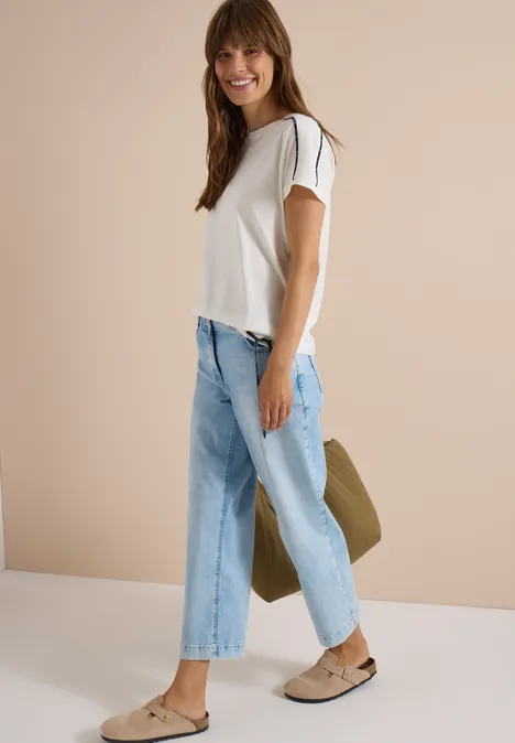 Jonge vrouw in wit T-shirt, lichtblauwe jeans en loafers, glimlachend met een tas in de hand.