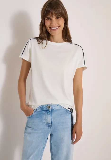 Een lachende vrouw draagt een wit t-shirt met schouderstrepen en een lichte jeans, met haar handen in de zakken.