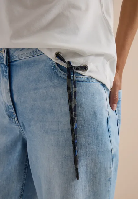 Persoon draagt lichtblauwe jeans met camouflage-trekkoord, hand in zak en wit T-shirt.