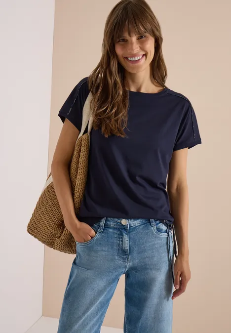 Jonge vrouw lacht in een casual outfit: donkerblauw t-shirt, spijkerbroek en rieten tas voor een neutrale achtergrond.