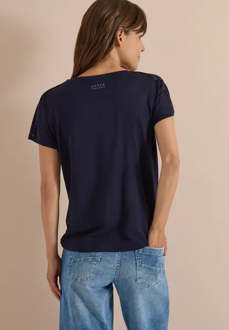 Rugaanzicht van een vrouw in een donkerblauw T-shirt en spijkerbroek tegen een neutrale achtergrond.