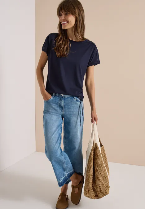 Vrouw in donkerblauw T-shirt, losse spijkerbroek, bruine mocassins en grote gevlochten tas op neutrale achtergrond.