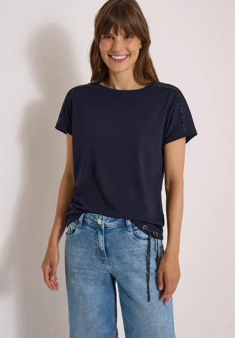Vrouw in marineblauw t-shirt met zijsamenvatting en blauwe jeans, lachend voor lichte achtergrond.
