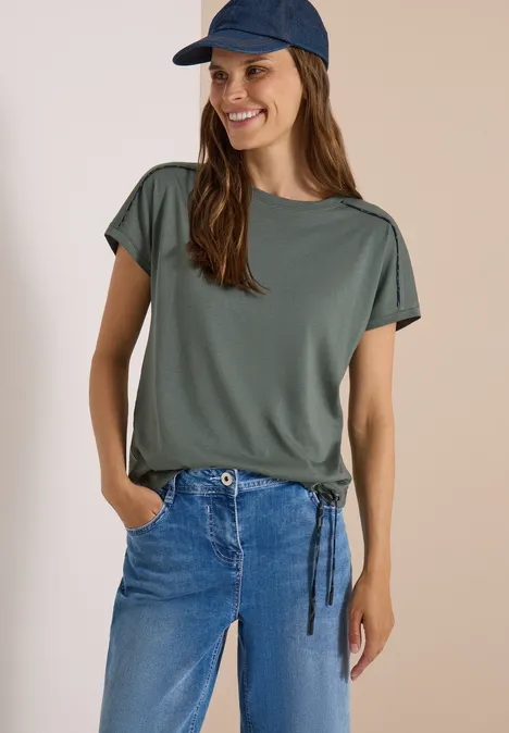 Vrouw in groen T-shirt, blauwe jeans en pet, glimlachend met handen in haar zakken.