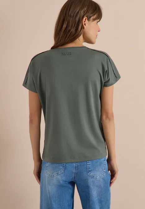 Vrouw in profielzicht, draagt groen T-shirt met korte mouwen en blauwe jeans tegen neutrale achtergrond.