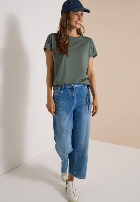 Jonge vrouw in groen T-shirt, wijde spijkerbroek, witte sneakers en pet tegen lichte achtergrond.