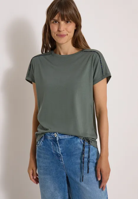 Vrouw met bruin haar draagt groen T-shirt met korte mouwen en koordjes, gecombineerd met blauwe jeans.