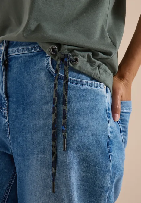 Close-up van een persoon met een groen shirt en losse blauwe spijkerbroek met trekkoord.