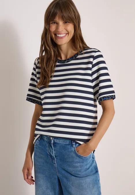 Frau mit braunen Haaren lächelnd, gestreiftes T-Shirt und Jeans vor hellem Hintergrund.