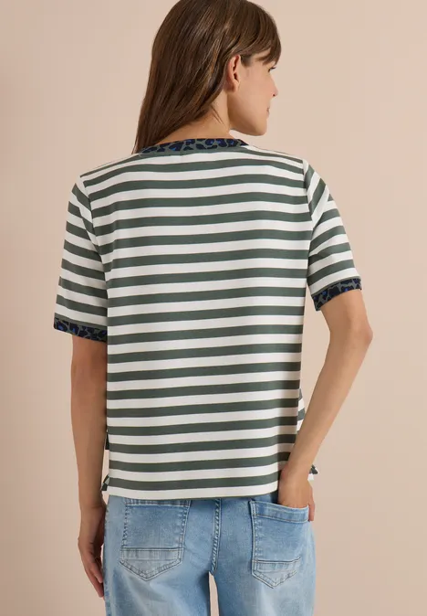 Eine Frau von hinten trägt ein grün-weiß gestreiftes Kurzarmshirt mit blauer Spitzenborte an Ärmeln und Saum und Jeans.