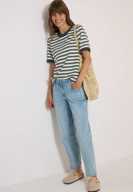 Frau mit gestreiftem Shirt, Jeans und Strohtasche lehnt an weißer Wand und lächelt.