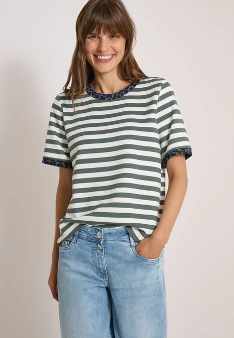 Eine lächelnde junge Frau in einem gestreiften T-Shirt und Jeans, eine Hand in der Hosentasche vor hellem Hintergrund.