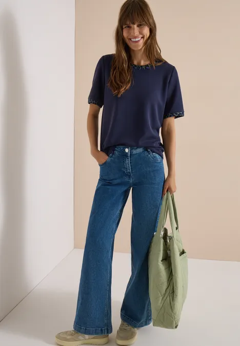 Eine lächelnde Frau trägt ein dunkelblaues T-Shirt, weite Jeans und hält eine grüne Tasche.