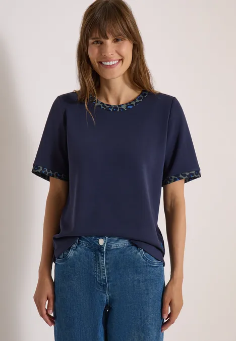 Frau in dunkelblauem T-Shirt mit gemustertem Saum und blauer Jeans, lächelnd