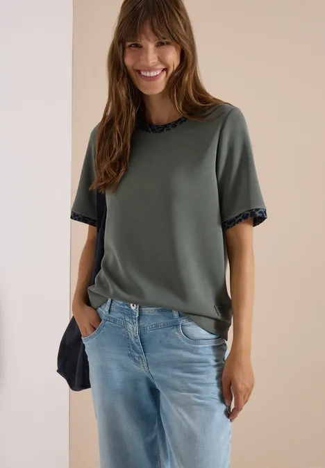 Een glimlachende vrouw draagt een groen T-shirt en een blauwe jeans.