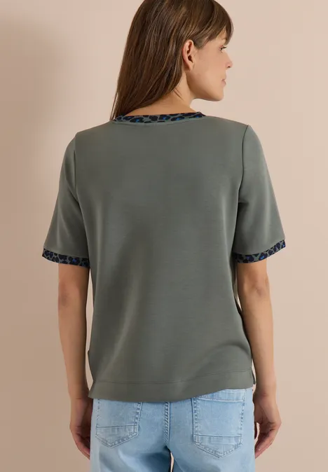 Vrouw van achter gefotografeerd in een grijs T-shirt met korte mouwen en ornamentale rand bij hals en mouwen.