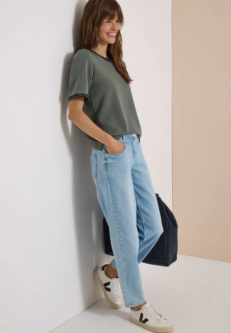 Vrolijke vrouw in groen T-shirt, lichtblauwe jeans en sneakers, leunend tegen muur met tas.