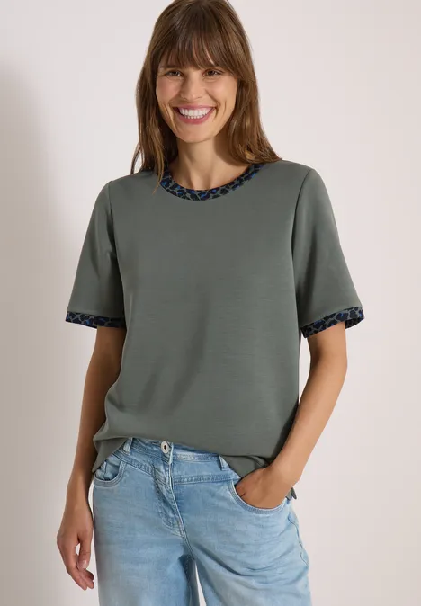 Lachende vrouw in kaki kort mouw top met patroonranden en lichtblauwe jeans.