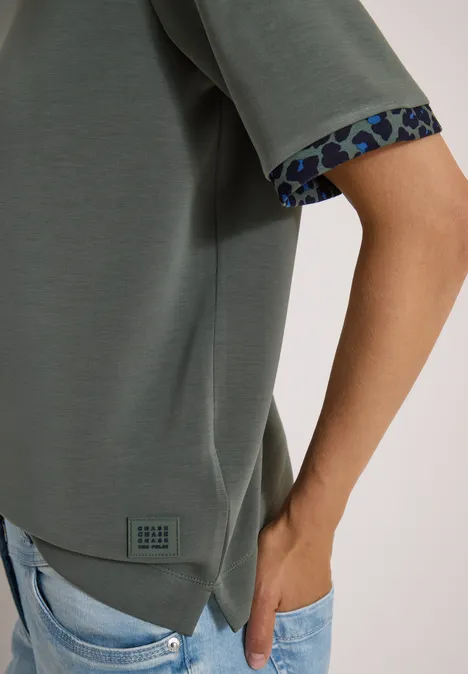 Olijfgroene top met gemêleerde mouwboord, lichtblauwe jeans, hand in de zak.