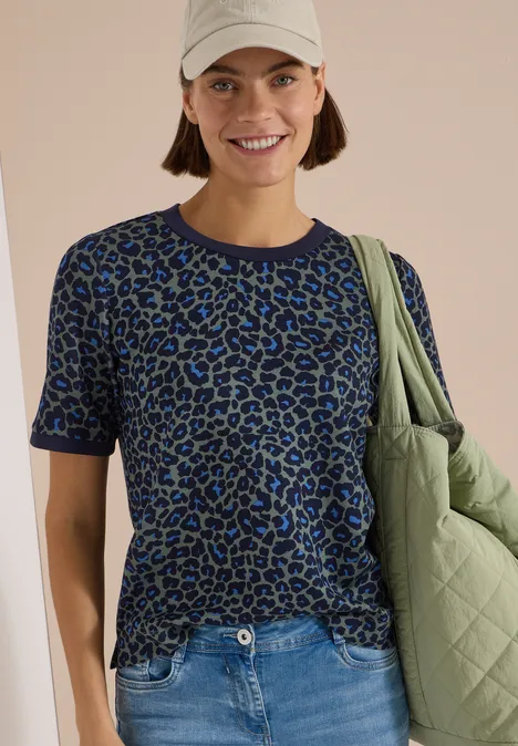 T-Shirt mit Rundhals und Leo-Muster dried olive green Frau lächelt in beiger Mütze, blau-grünem Leo-Shirt, Jeans und grünem Steppbeutel.