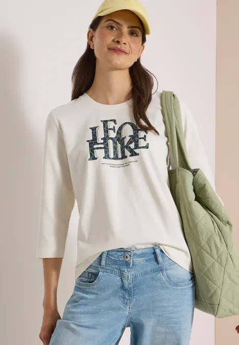 T-shirt à manches 3/4 avec inscription, style léopard vanilla white Frau in Jeans, weißem 3/4-Shirt mit 'HERO HIKE'-Schriftzug, gelber Mütze und grüner, gesteppter Tasche.