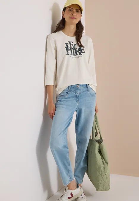 T-shirt à manches 3/4 avec inscription, style léopard vanilla white Junge Frau in weißem Shirt, hellen Jeans und gelber Baseballkappe, hält eine grüne Tasche.