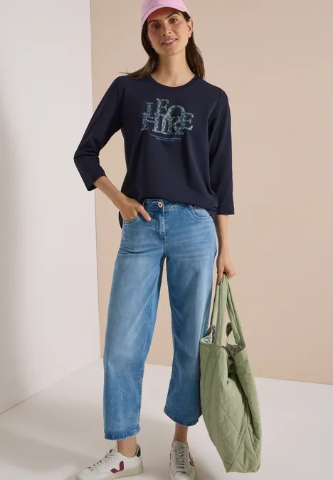 T-shirt à manches 3/4 avec inscription, style léopard urban dark blue Frau mit dunkelblauem Shirt, weiten Jeans, Baseballcap und großer grüner Tasche vor hellem Hintergrund