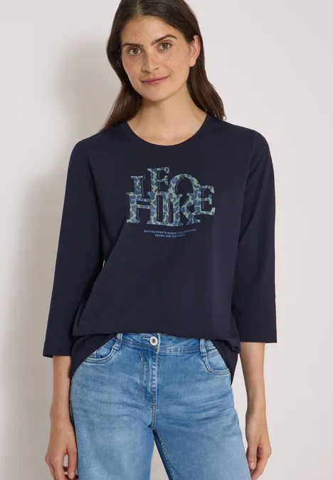 T-shirt à manches 3/4 avec inscription, style léopard urban dark blue Eine Frau in Jeans und dunkelblauem Langarmshirt mit Aufdruck steht vor hellem Hintergrund.