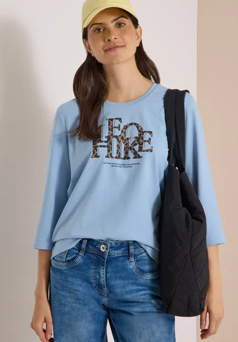 T-shirt à manches 3/4 avec inscription, style léopard easy blue Frau mit beiger Kappe, hellblauem 3/4-Shirt mit Leo-Print-Schriftzug, Jeans und schwarzer Tasche vor neutralem Hintergrund.