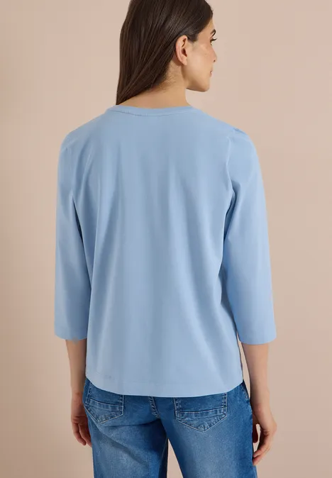 T-shirt à manches 3/4 avec inscription, style léopard easy blue Frau von hinten in lockerem hellblauen 3/4-Arm-Shirt und Jeans vor neutralem Hintergrund