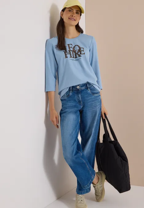 T-shirt à manches 3/4 avec inscription, style léopard easy blue Frau in gelber Baseballmütze, hellblauem Langarmshirt mit Aufdruck, Jeans und schwarzer Tasche.