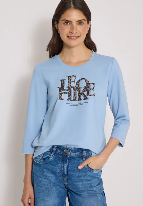 T-shirt à manches 3/4 avec inscription, style léopard easy blue Frau trägt blaues 3/4-Arm-Shirt mit 'FOR HIKE'-Aufdruck und Jeans, lächelnd vor neutralem Hintergrund