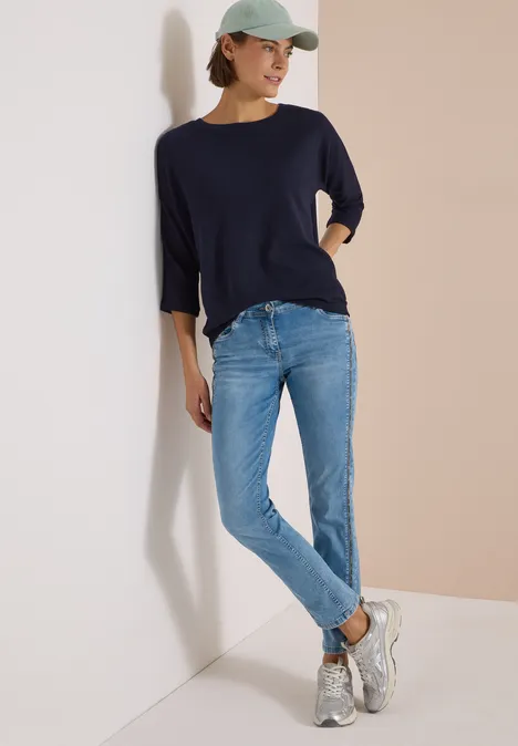 3/4-Arm Shirt mit Waffelstruktur urban dark blue Frau mit Cap, dunkelblauem 3/4-Shirt, hellen Jeans und Sneakers lehnt entspannt an einer Wand.