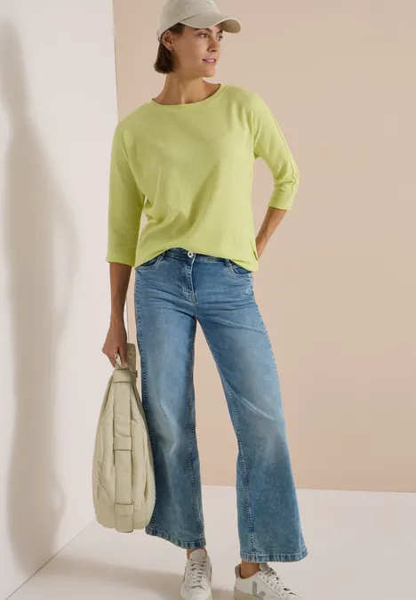 3/4-Arm Shirt mit Waffelstruktur citrin green Eine Frau lehnt an der Wand, trägt hellgrünen Pullover, Jeans, Cap und Tasche.