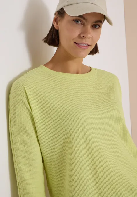 3/4-Arm Shirt mit Waffelstruktur citrin green Junge Frau mit beiger Baseballcap und hellgrünem Pullover lehnt lächelnd an heller Wand.