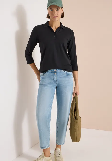Person steht an Wand, trägt schwarzes Dreiviertelärmel-Shirt, hellblaue Jeans und hält einen olivfarbenen Stoffbeutel.
