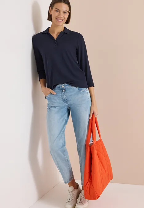 Eine Person trägt ein dunkelblaues Langarmshirt, helle Jeans, weiße Sneakers und hält eine orange Tasche.