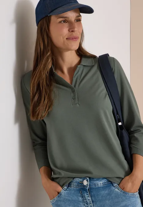 Polo à manches 3/4 avec boutons décoratifs dried olive green Frau lehnt an der Wand, trägt olivgrünes Poloshirt mit 3/4-Ärmeln, Jeans, blaue Cap und Schultertasche.