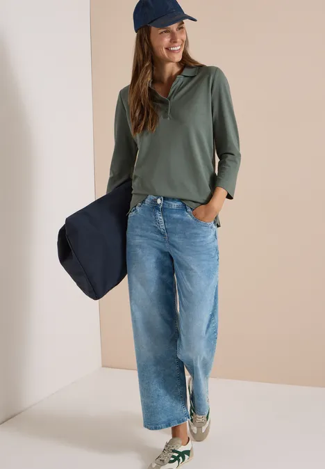 Polo à manches 3/4 avec boutons décoratifs dried olive green Frau mit blauer Jeans, olivgrünem Shirt und Cap hält navy Jacke, lässiger Alltagslook.