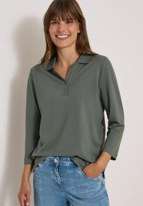 Polo à manches 3/4 avec boutons décoratifs dried olive green Frau mit langärmligem grünen Poloshirt, Jeans, Hände in den Taschen, lächelnd vor weißem Hintergrund.