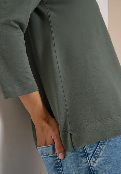 Polo à manches 3/4 avec boutons décoratifs dried olive green Person lehnt an Wand, trägt grünes Shirt mit 3/4-Ärmeln und blaue Jeans, Hand steckt in Tasche.