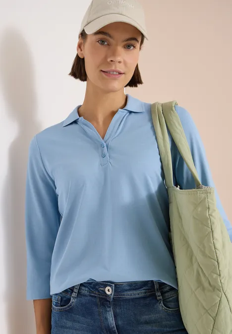 Polo à manches 3/4 avec boutons décoratifs easy blue Eine Person trägt ein hellblaues Langarmshirt, Jeans und eine grüne, gesteppte Tragetasche.