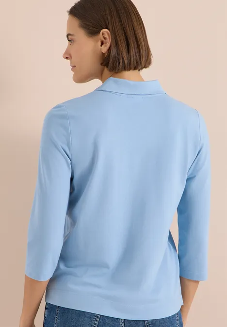 Polo à manches 3/4 avec boutons décoratifs easy blue Frau mit dunklem Bob trägt hellblaues 3/4-Arm-Shirt, steht blickabgewandt vor neutralem Hintergrund.