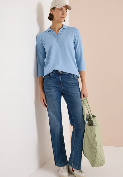 Polo à manches 3/4 avec boutons décoratifs easy blue Frau trägt hellblaues Poloshirt, weite Jeans und hält eine grüne Stofftasche vor zweifarbigem Hintergrund.